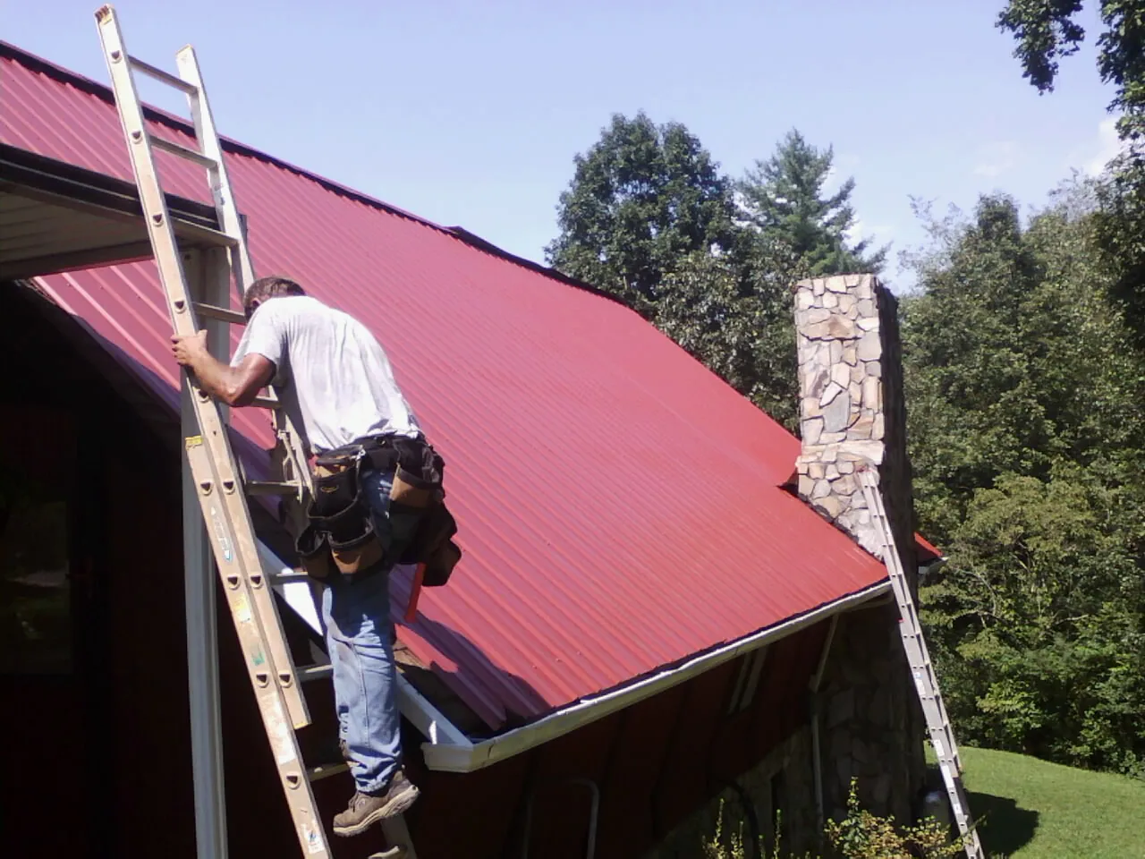 Roofer on a ladder beside a red metal roof and stone chimney — Soffit & Fascia Repair by Roofing Brothers in Millers Creek